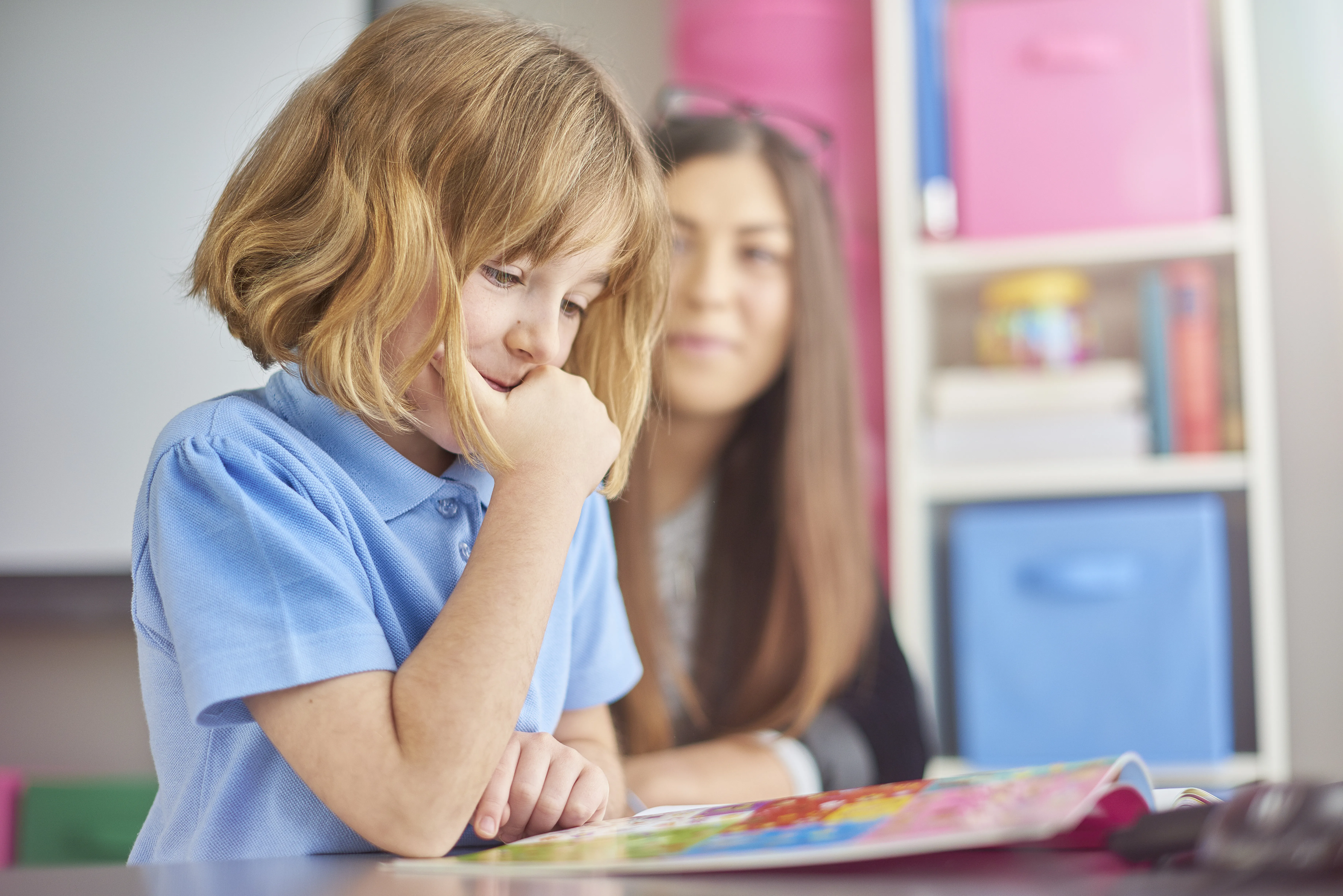a primary school girl is sitting at her desk looking at her book and thinking . Her young teacher looks on  in the background giving support .